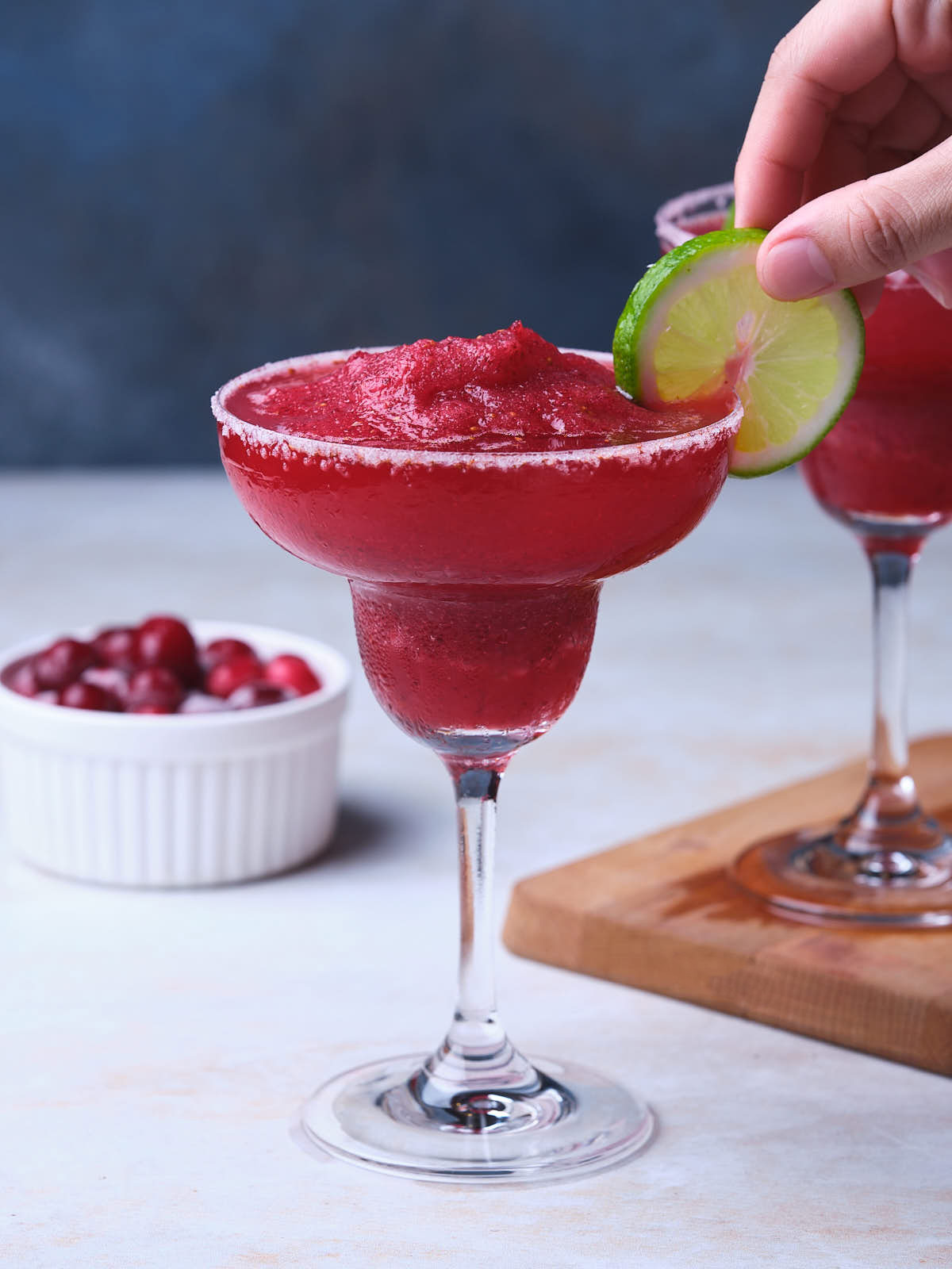 A hand garnishes a frozen cranberry margarita glass with a slice of lime.