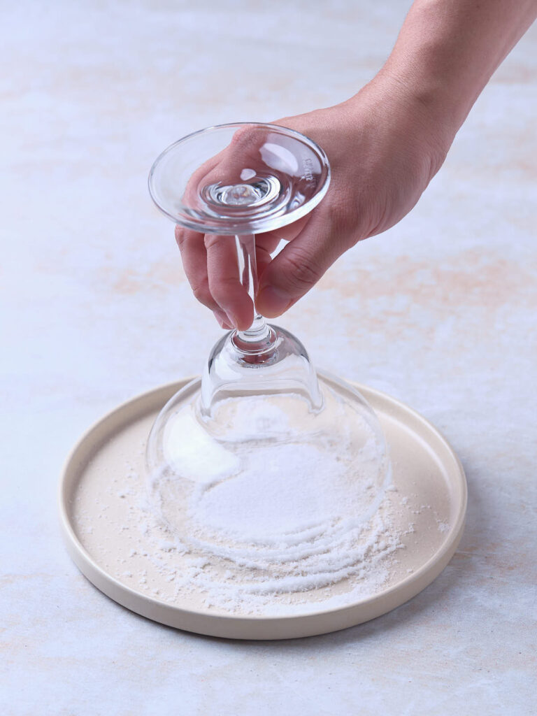 A hand dipping the rim of an upside-down wine glass into a plate of salt.