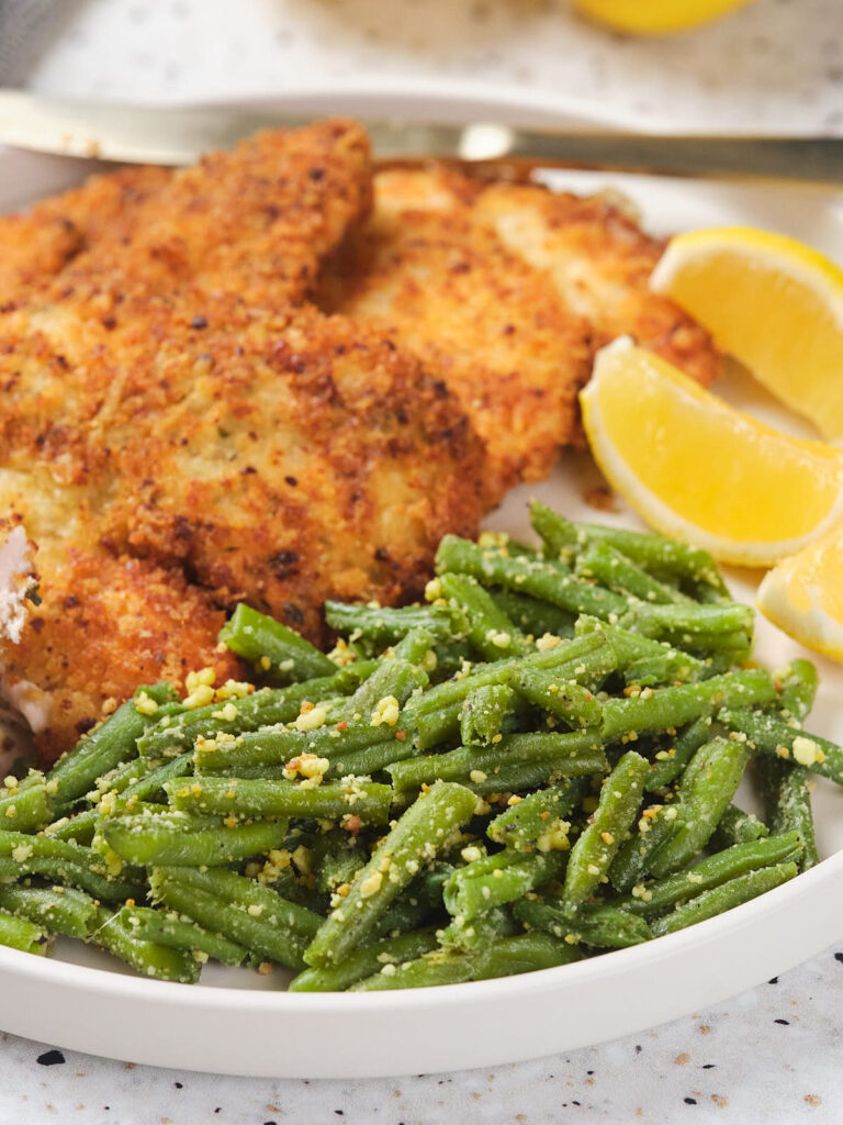 A plate with chicken cutlets, frozen roasted green beans topped with seasoning, and lemon wedges.