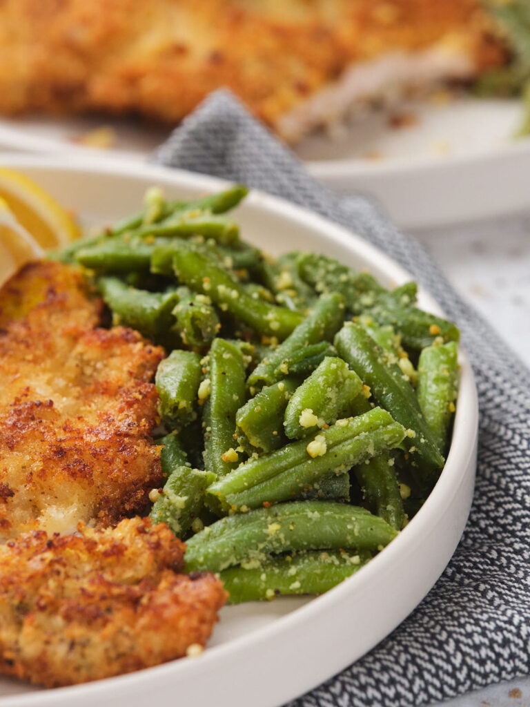 A plate of breaded chicken and seasoned green beans on a white dish placed on a gray napkin.