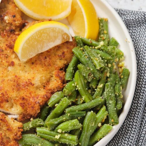 Plate with breaded chicken, lemon wedges, and green beans topped with grated cheese.