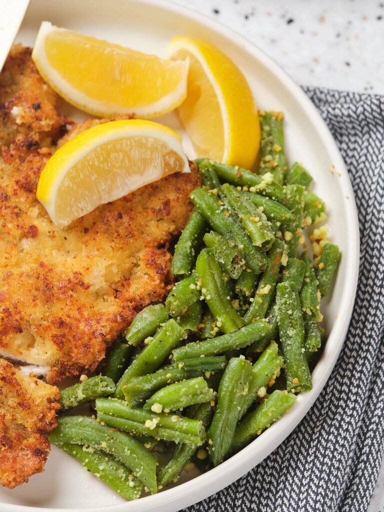 Plate with breaded chicken, lemon wedges, and green beans topped with grated cheese.