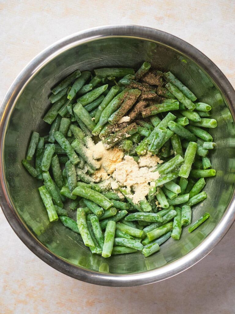 A metal bowl containing cut green beans seasoned with pepper, salt, and garlic powder.