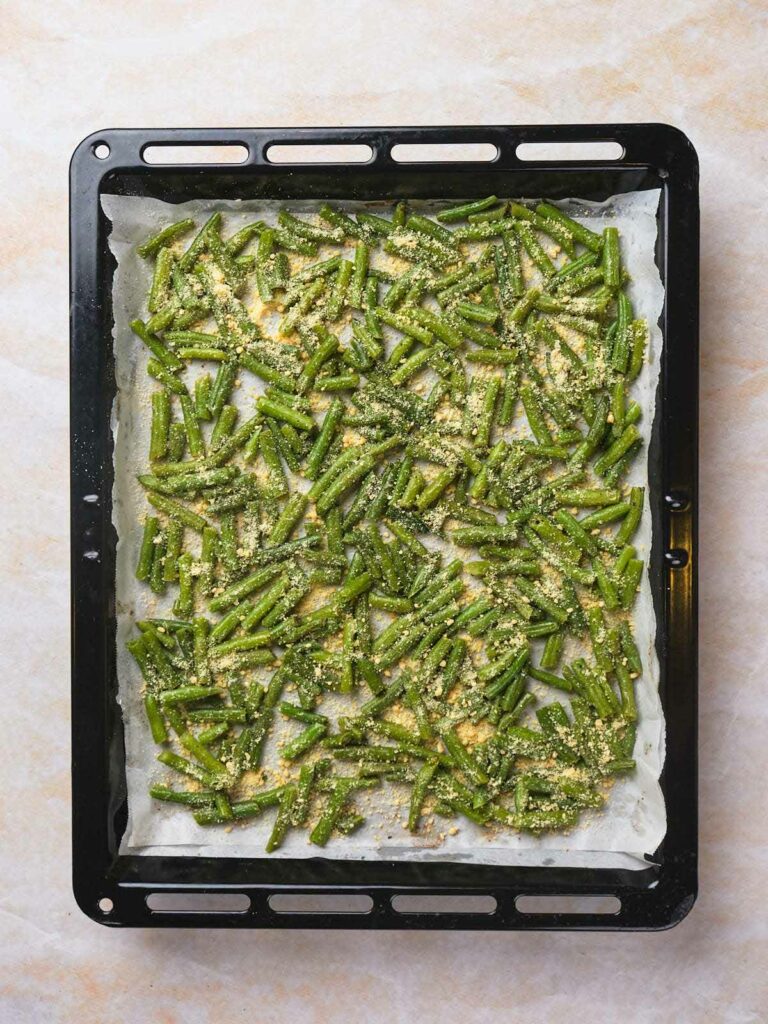 Baking tray with sliced green beans coated in breadcrumbs and cheese on parchment paper.