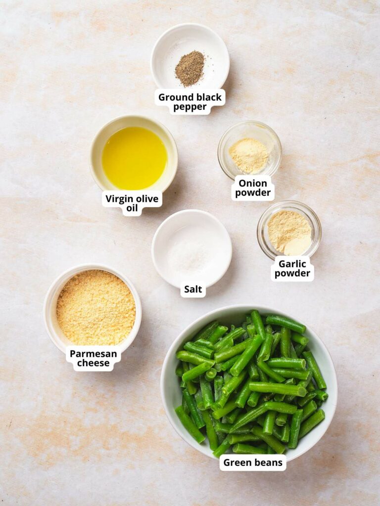 Ingredients for frozen roasted green beans arranged on a surface: green beans, Parmesan cheese, virgin olive oil, ground black pepper, salt, onion powder, garlic powder.