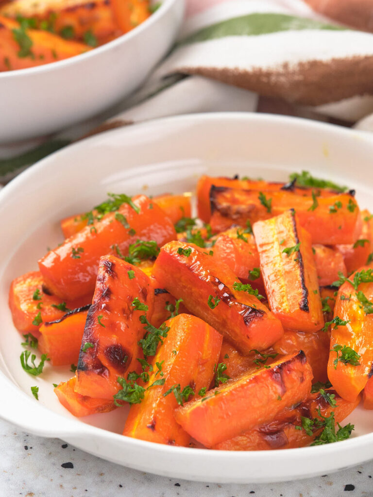 A white bowl filled with roasted carrot sticks, garnished with chopped parsley, placed on a light-colored table.