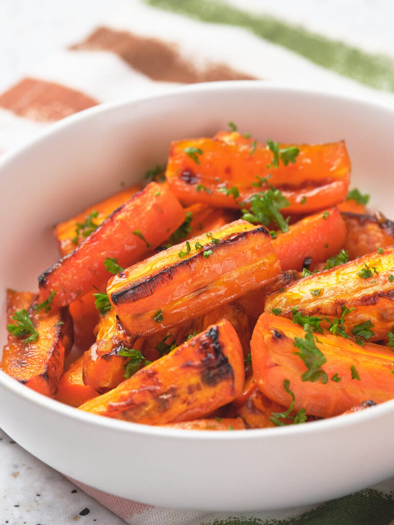 A white bowl filled with honey glazed carrots garnished with chopped parsley on a striped cloth.