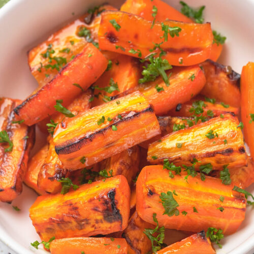 A bowl of honey glazed carrots garnished with chopped parsley on a white surface.