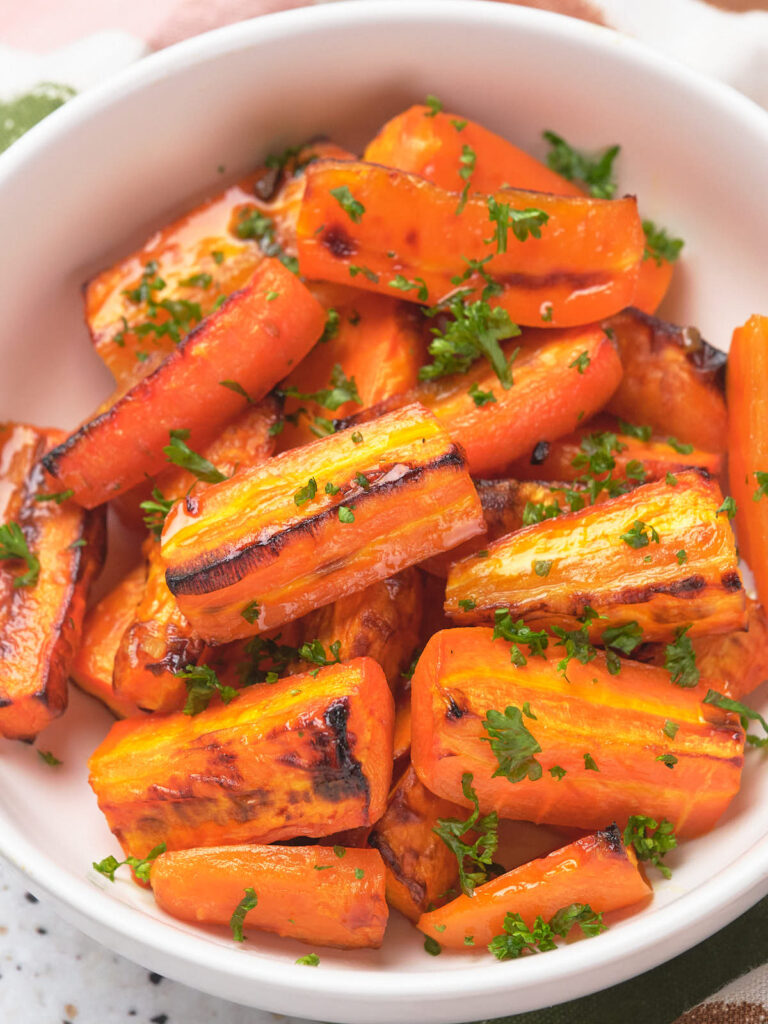 A bowl of honey glazed carrots garnished with chopped parsley on a white surface.