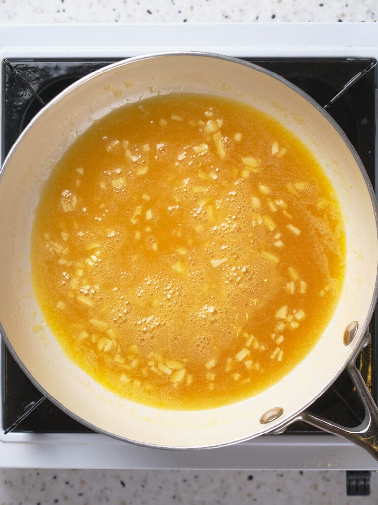 A frying pan on a stovetop contains a bubbling melted butter with chopped garlic pieces.