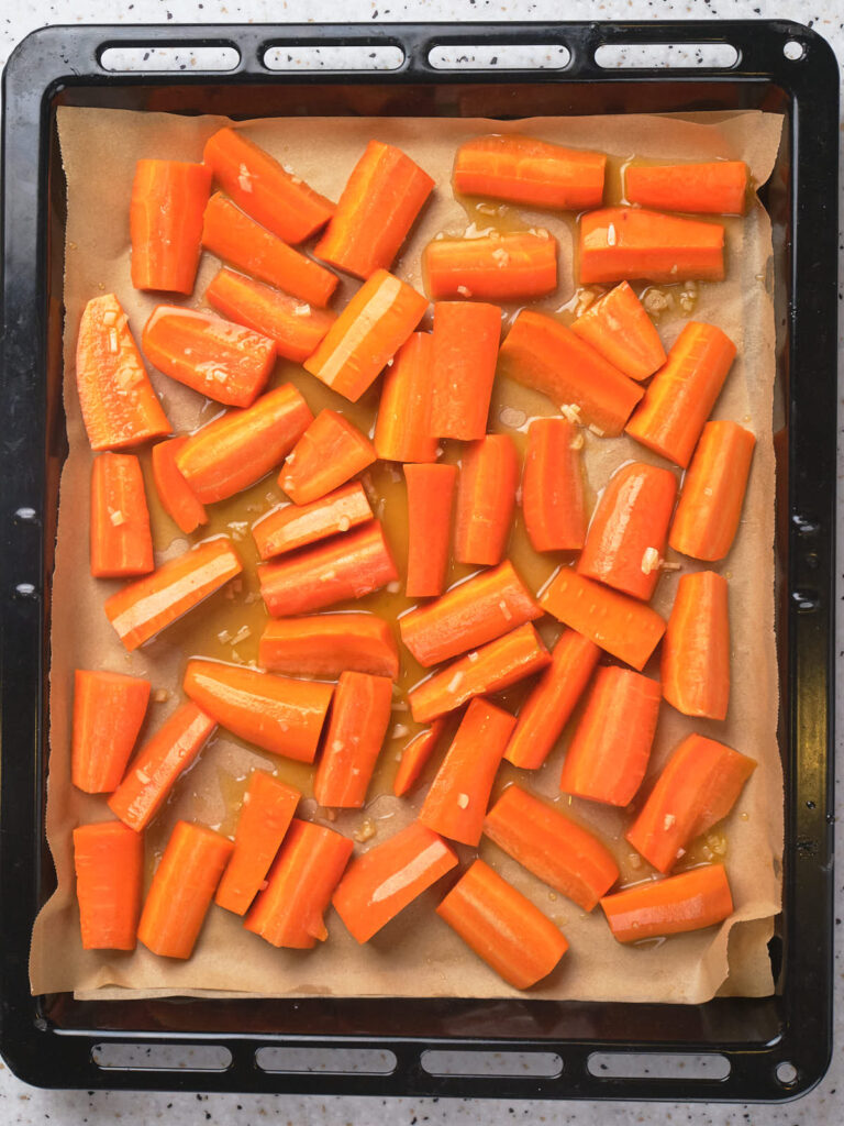 Chopped carrots tossed in oil on a parchment-lined baking tray, ready for roasting.