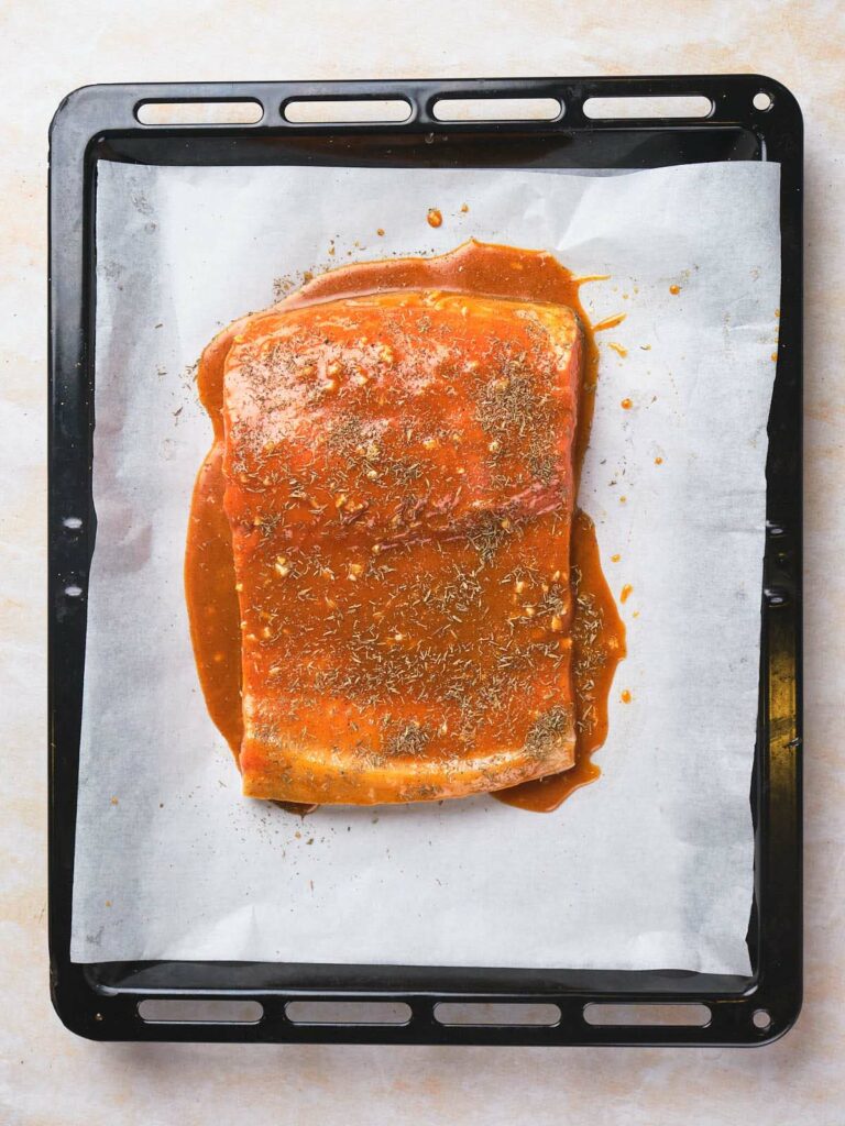 Raw, marinated salmon fillet on a parchment-lined baking sheet, seasoned with herbs and a reddish sauce.