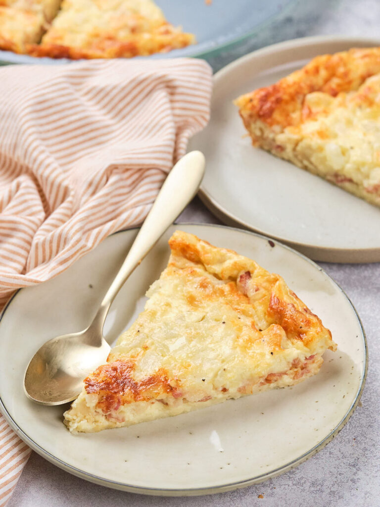 A slice of quiche on a plate with a spoon next to it, accompanied by a striped cloth in the background.