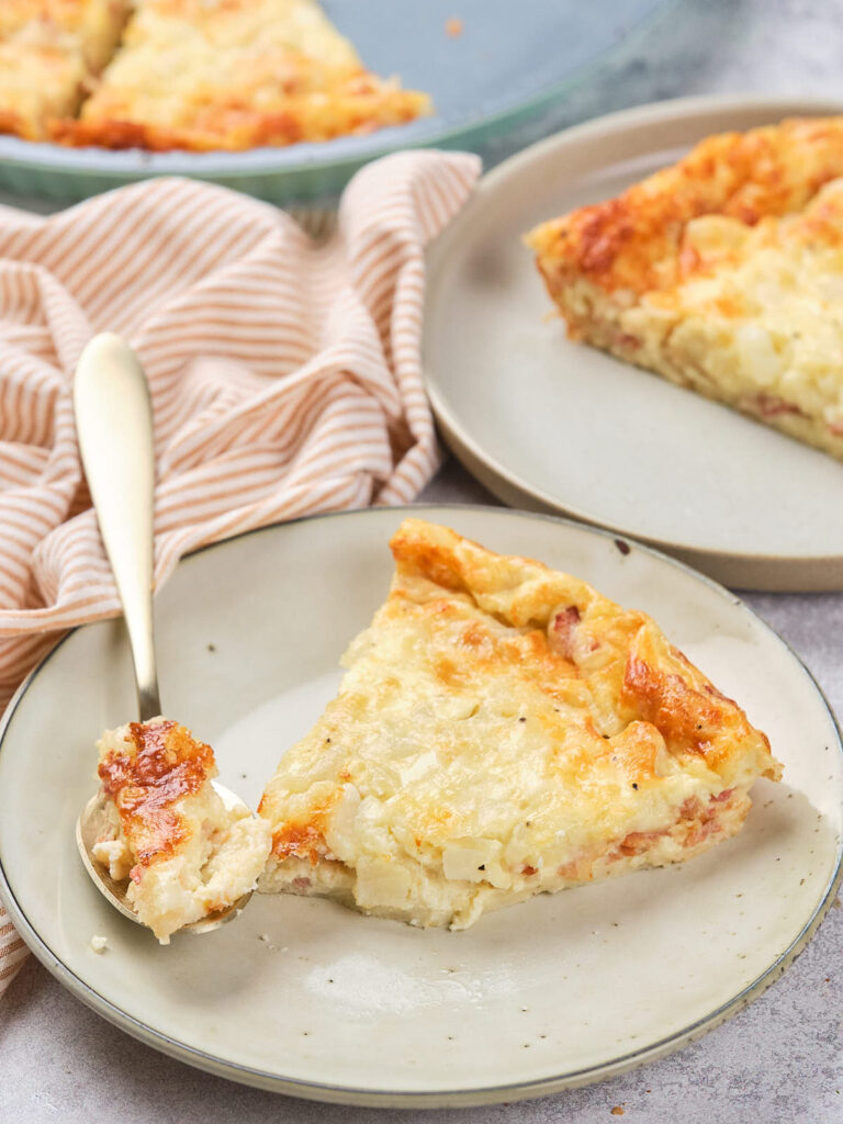 Slice of quiche on a plate with a fork, a striped cloth, and another pie slice in the background.