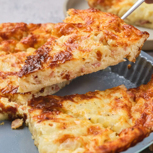 A spatula lifts a slice of cheesy quiche from a pie dish.