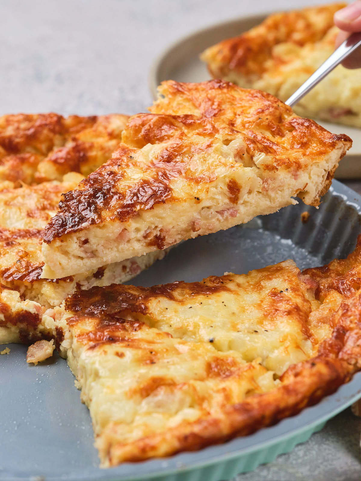 A spatula lifts a slice of cheesy quiche from a pie dish.