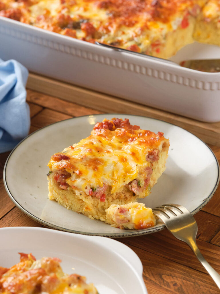 A slice of cheesy casserole with vegetables and meat on a plate, with a fork beside it. A large dish containing more casserole is visible in the background.