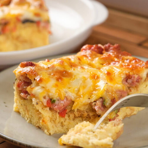 A slice of cheesy breakfast casserole with sausage and peppers on a plate, partially cut with a fork.