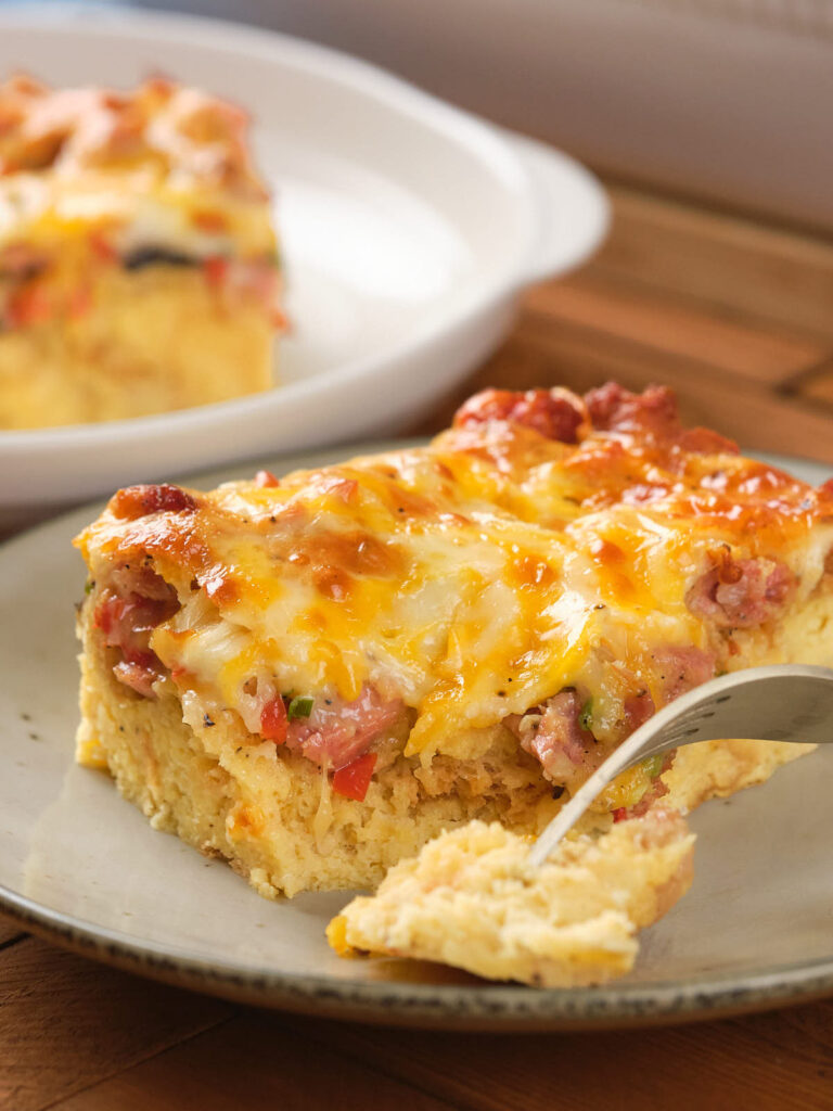 A slice of cheesy breakfast casserole with sausage and peppers on a plate, partially cut with a fork.