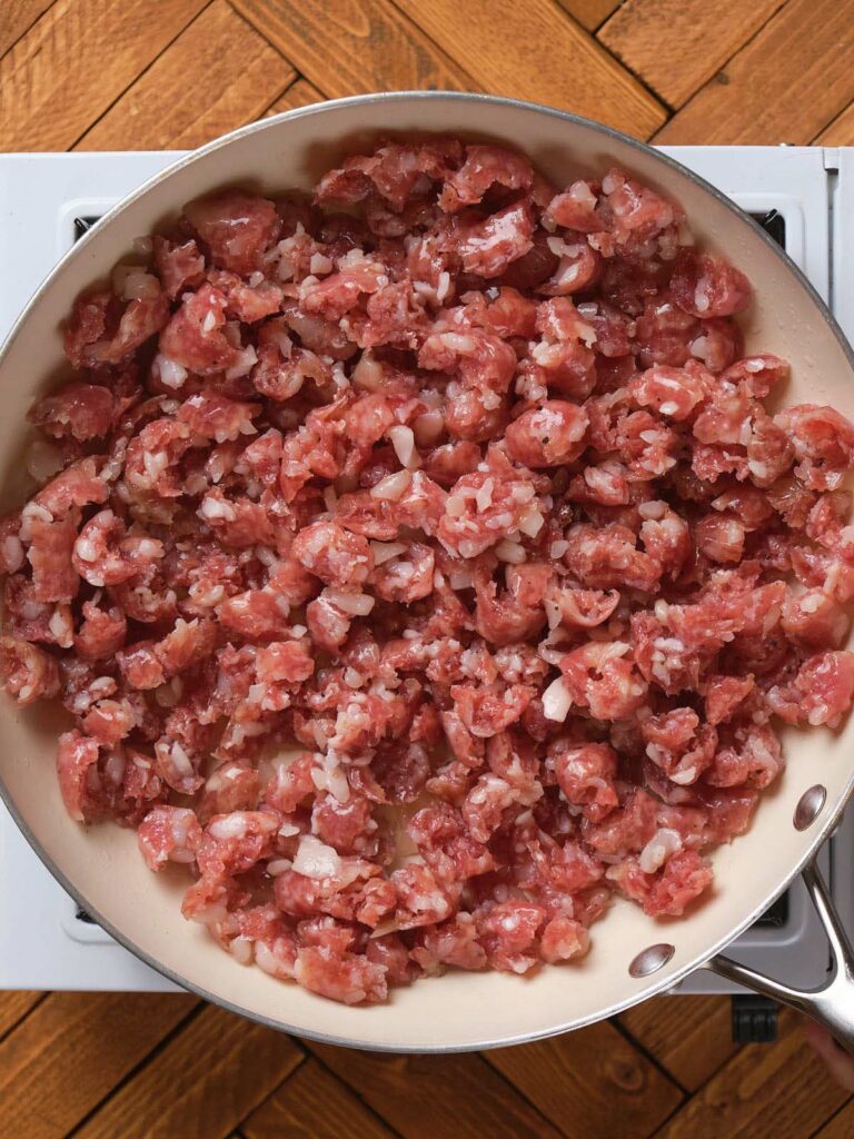 A frying pan filled with raw, diced meat on a stove, placed against a wooden tile background.