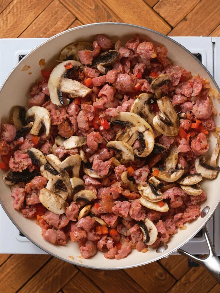 A skillet on a stove contains cooked ground meat, sliced mushrooms, and small diced red vegetables.