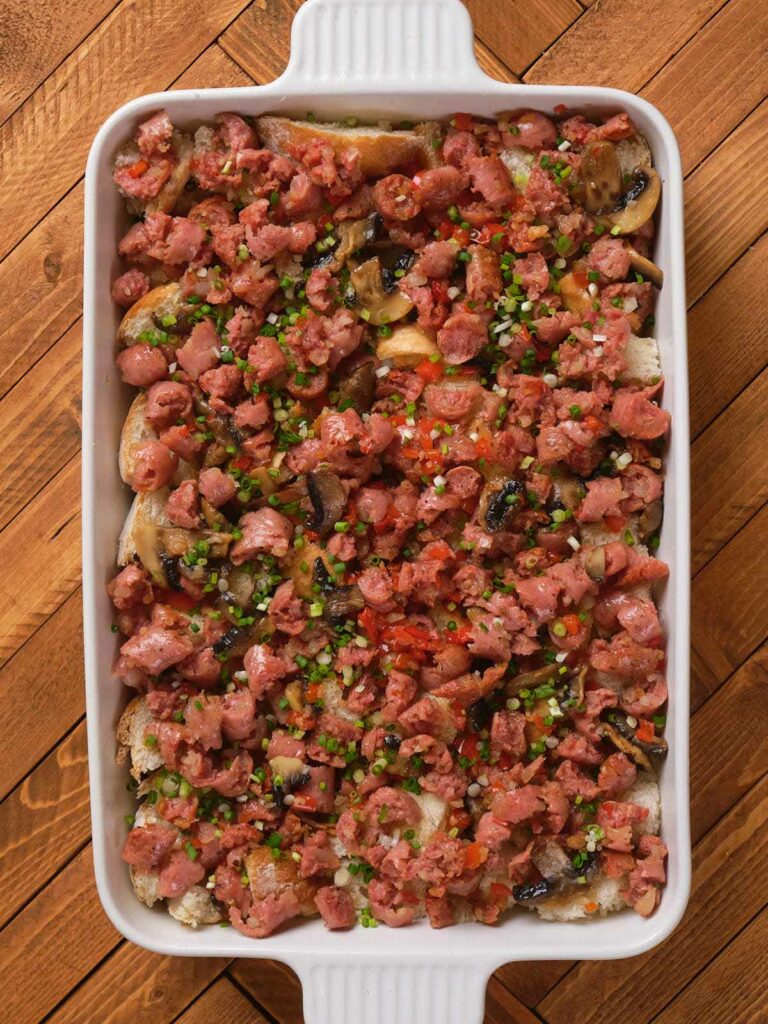 A baking dish filled with chopped sausage, mushrooms, and herbs, placed on a wooden surface.