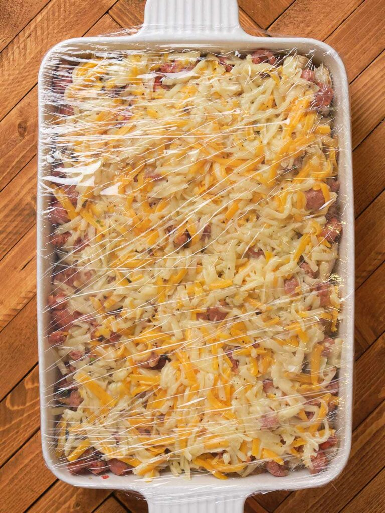 A casserole dish with shredded cheese on top, covered in plastic wrap, placed on a wooden surface.