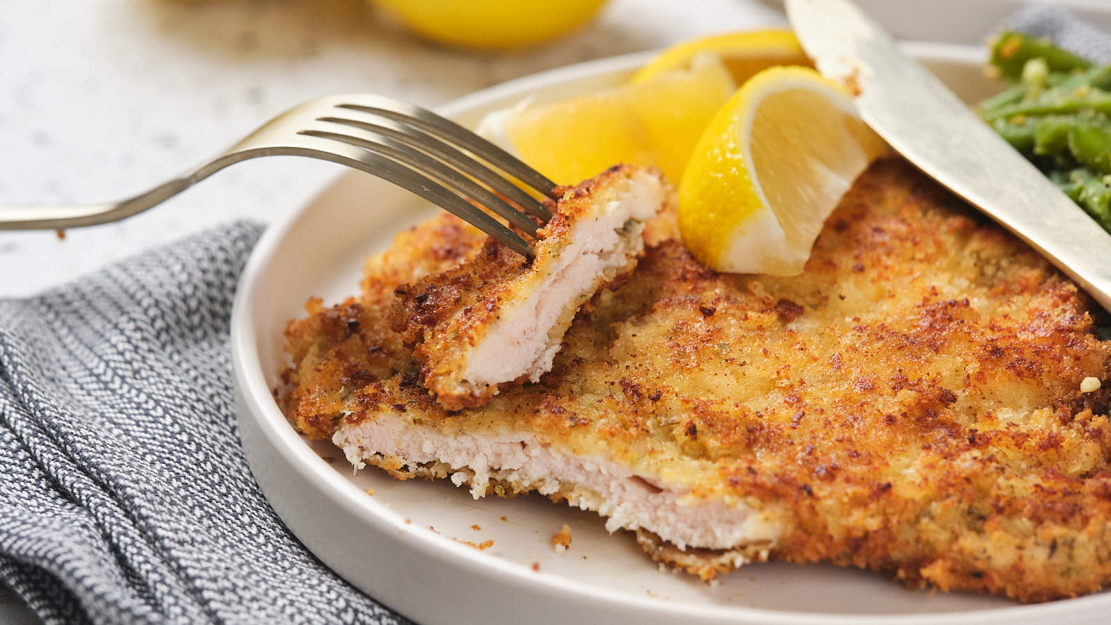 Piece of chicken cutlet on a fork resting over a larger portion with a lemon wedge.