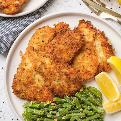 Parmesan crusted chicken on a white plate garnished with beans and lemon slices.