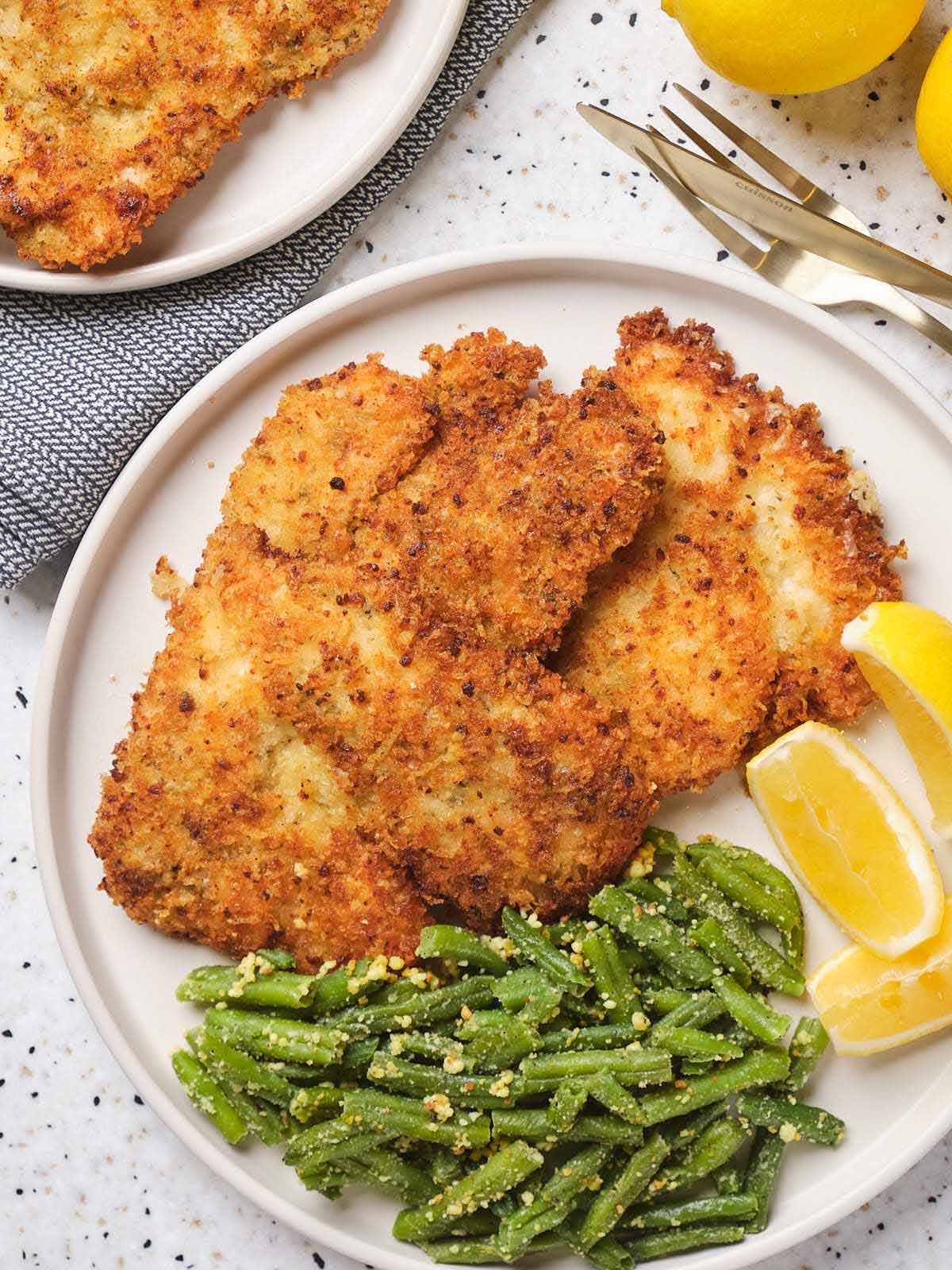 Parmesan crusted chicken on a white plate garnished with beans and lemon slices.