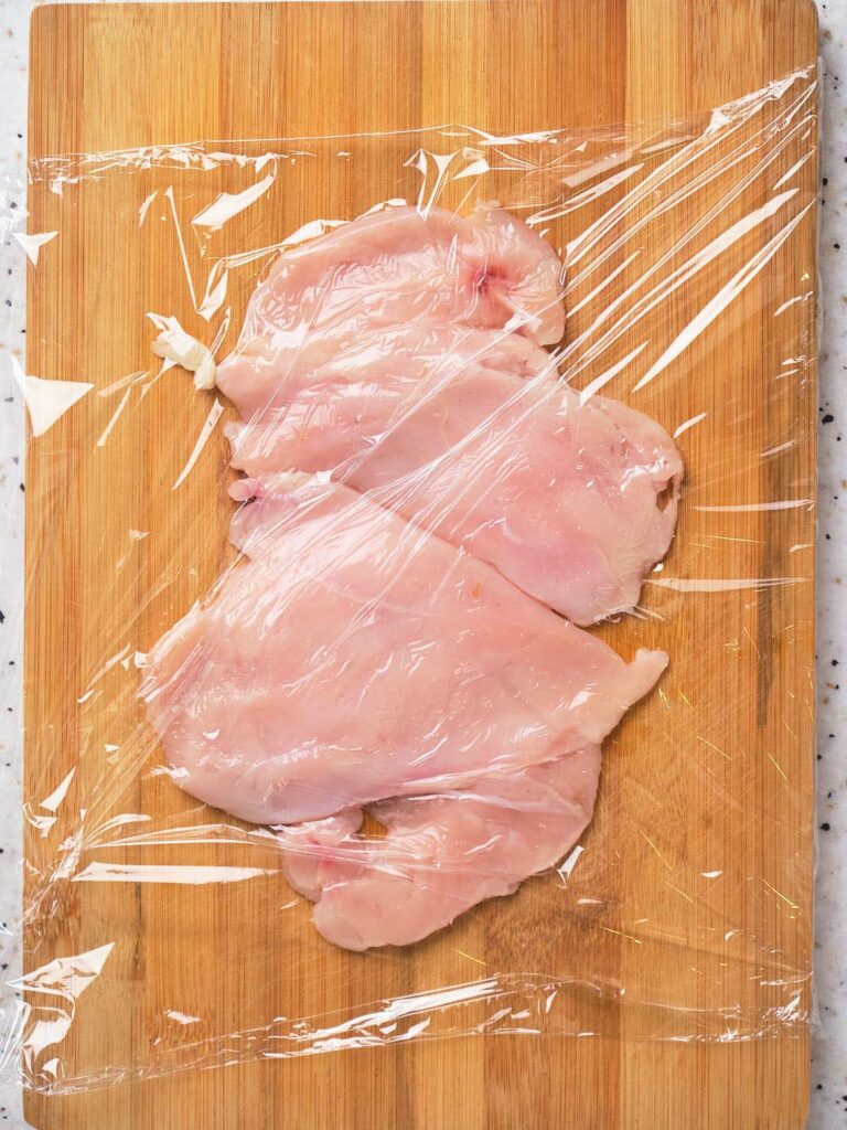 Raw chicken on a cutting board and covered with plastic wrap.