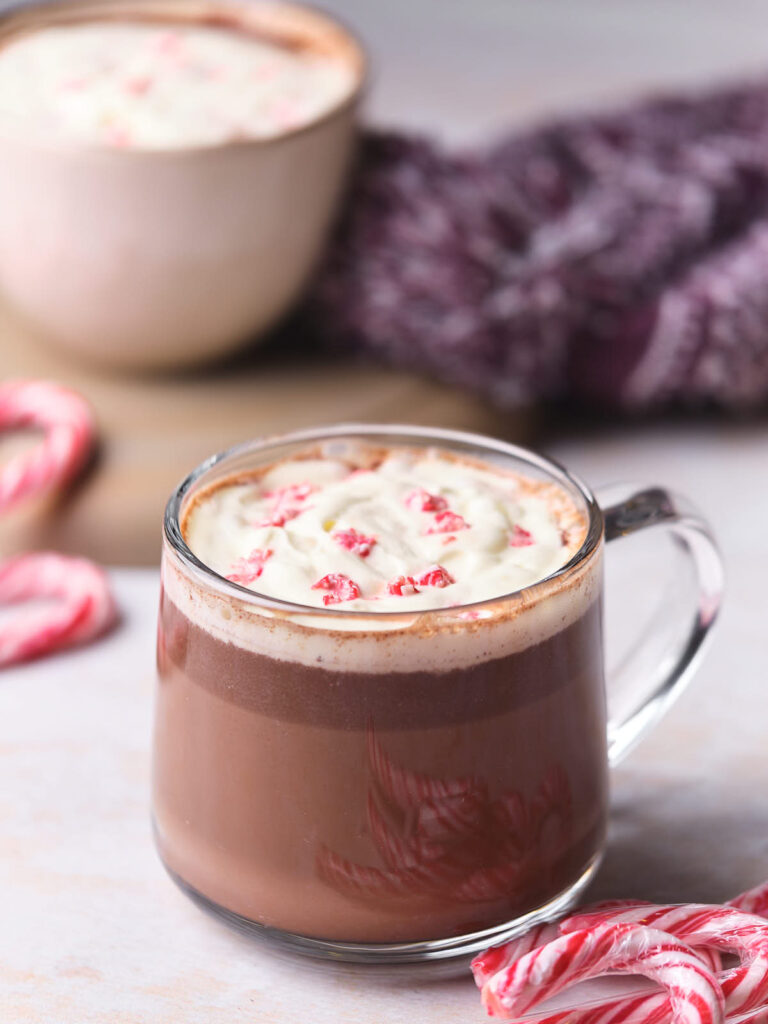 A glass cup of hot chocolate topped with whipped cream and crushed peppermint candy, next to candy canes, with another cup and a purple cloth in the background.