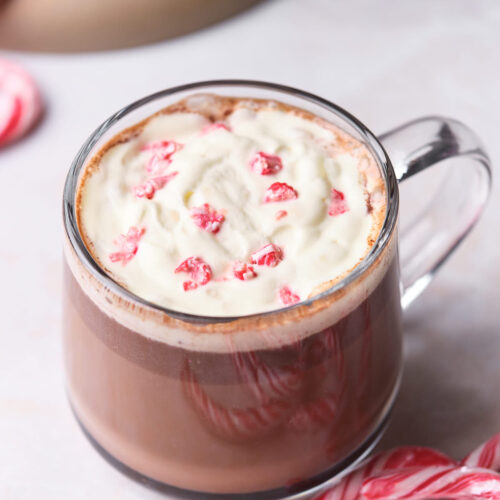 A glass mug of hot chocolate topped with whipped cream and crushed candy canes, next to whole candy canes on a light surface.