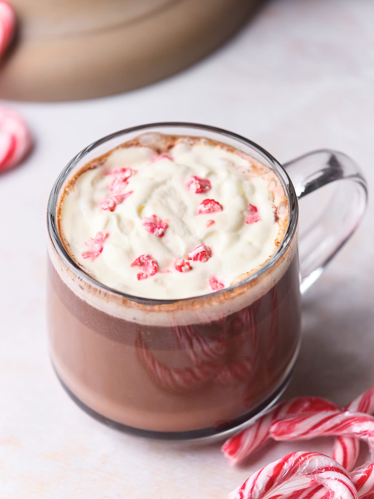 A glass mug of hot chocolate topped with whipped cream and crushed candy canes, next to whole candy canes on a light surface.