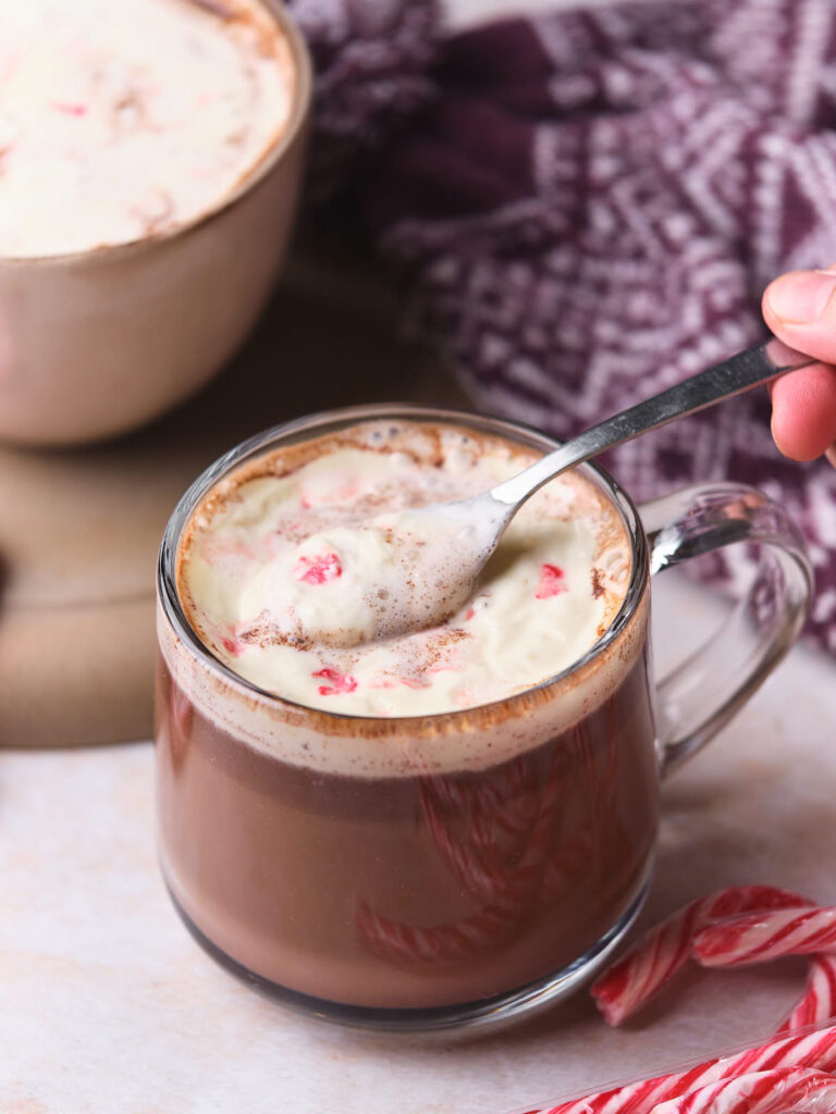 A clear mug of hot chocolate topped with whipped cream being stirred by a hand holding a spoon. Peppermint pieces are visible, and a candy cane is beside the mug. A knitted blanket is in the background.