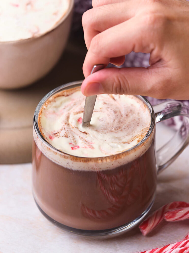 A hand stirs whipped cream into a mug of hot cocoa with a spoon, next to candy canes.