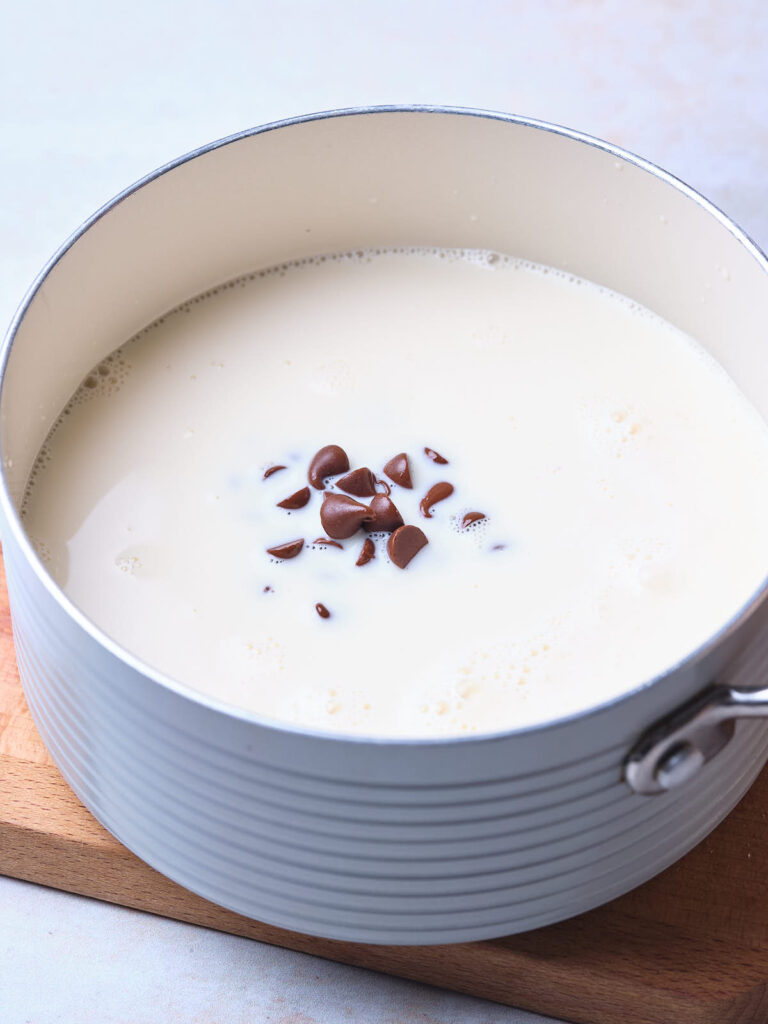 A saucepan with milk and melting chocolate chips inside, placed on a wooden board.