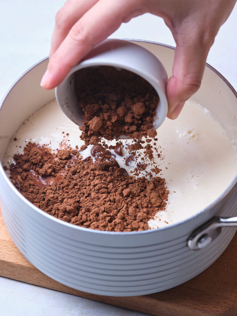 A hand pours cocoa powder from a bowl into a pot of milk on a wooden board.