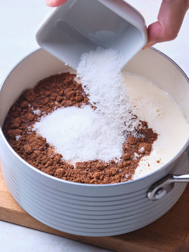 Pouring sugar into a pot with cocoa powder and milk.