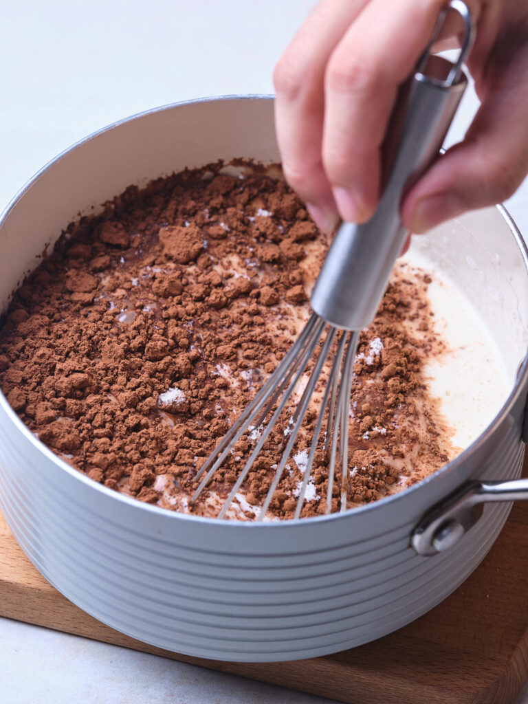 A hand whisking cocoa powder and milk in a gray pot.