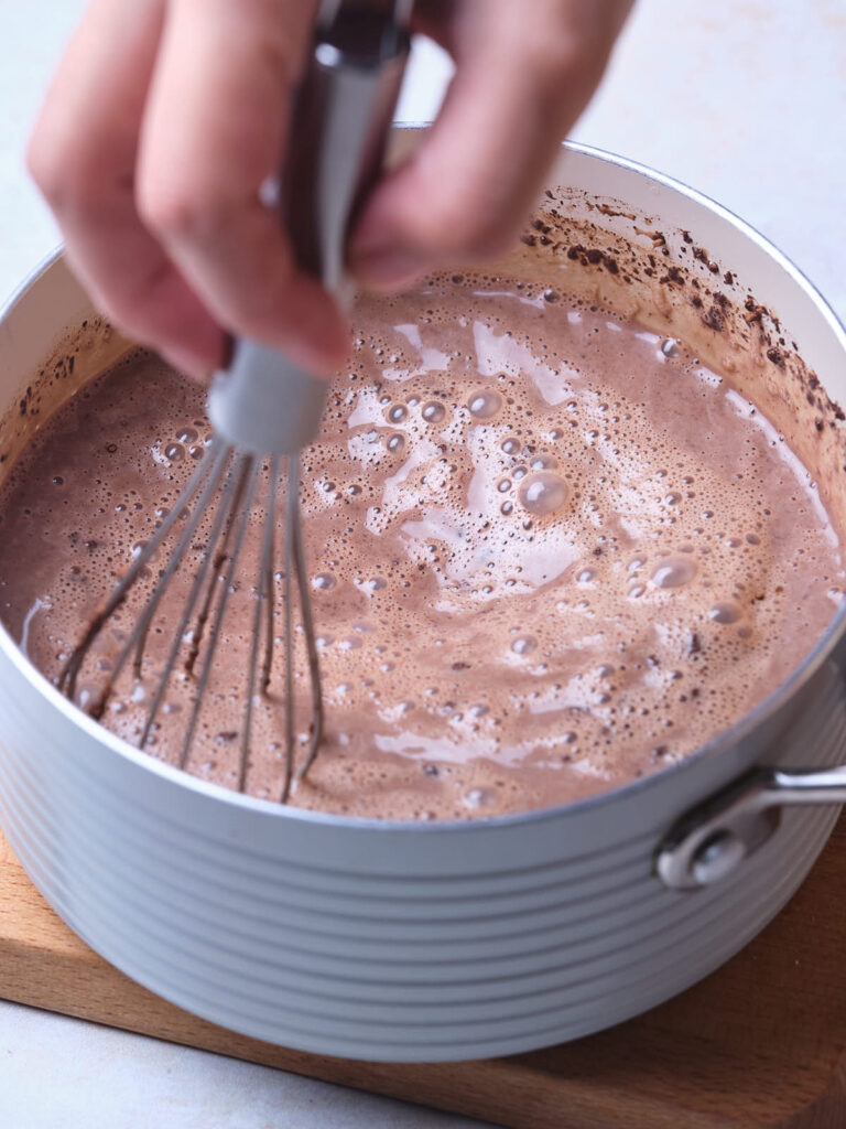 Hand whisking frothy chocolate mixture in a pot on a wooden board.