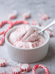 A bowl of peppermint ice cream with a spoon, surrounded by peppermint candies on a gray surface.