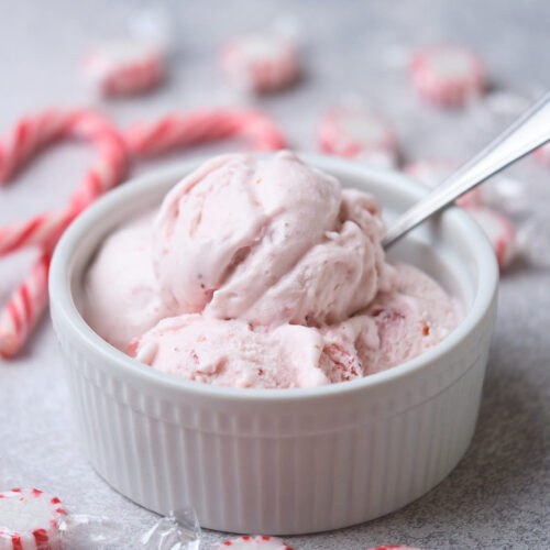 A bowl of peppermint ice cream with a spoon, surrounded by peppermint candies on a gray surface.