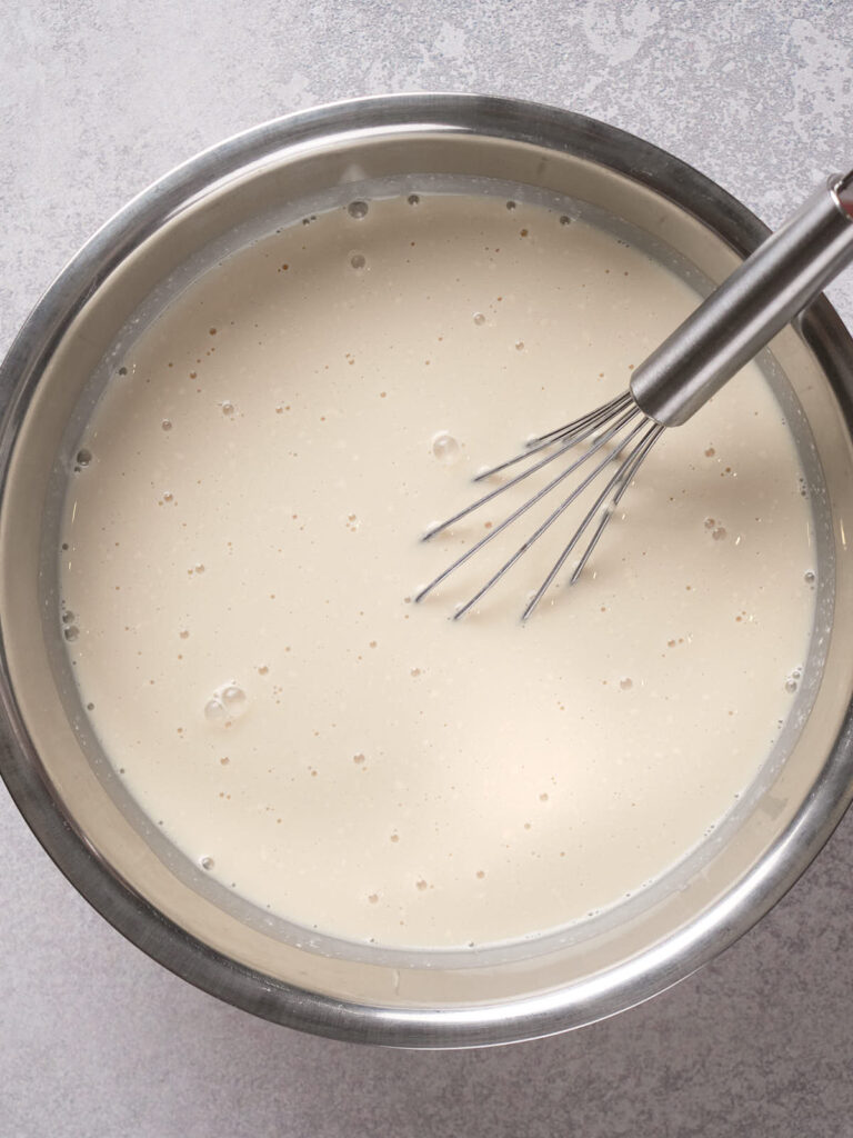 A stainless steel bowl filled with a light beige batter and a whisk resting inside.