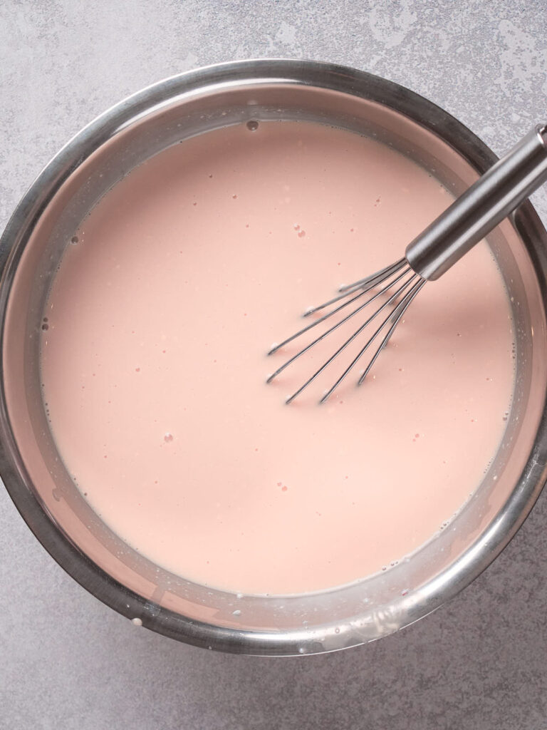 A stainless steel bowl with a whisk and light pink liquid inside, placed on a gray surface.