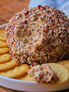 Pineapple cheese ball coated with chopped nuts, served on a white plate with round crackers.