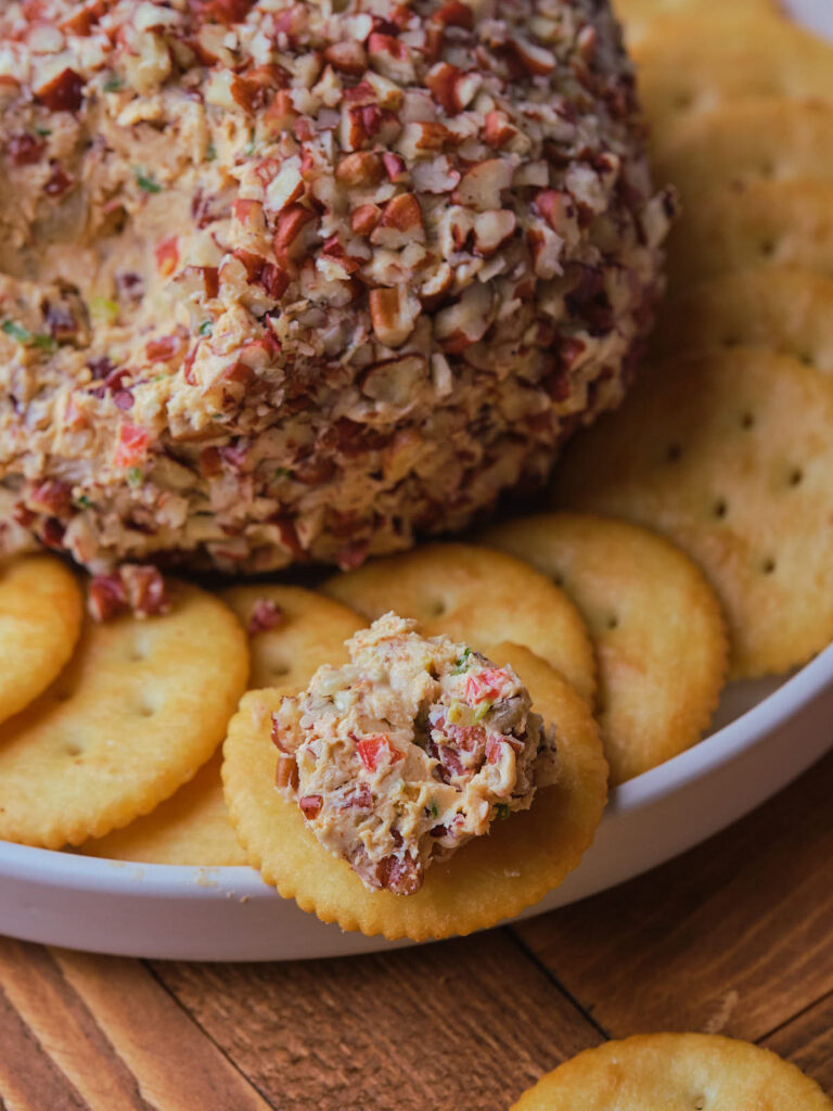 Pineapple cheese ball coated with chopped nuts is placed on a white plate surrounded by round crackers.