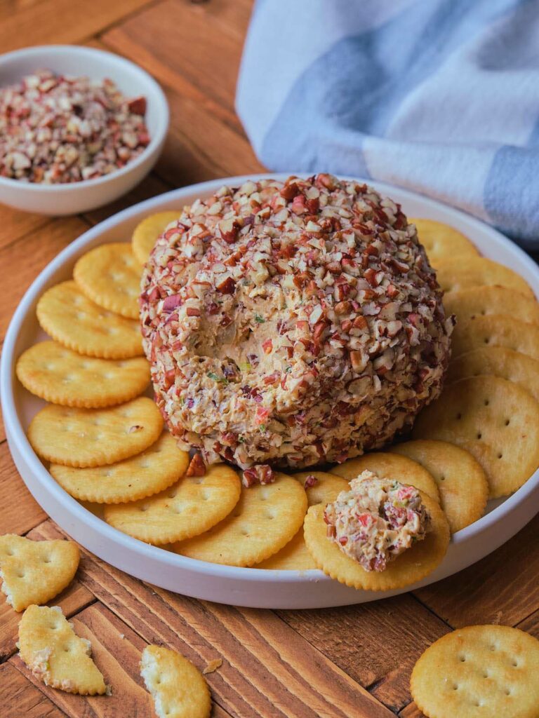 A round pineapple cheese ball coated in crushed nuts on a white plate.