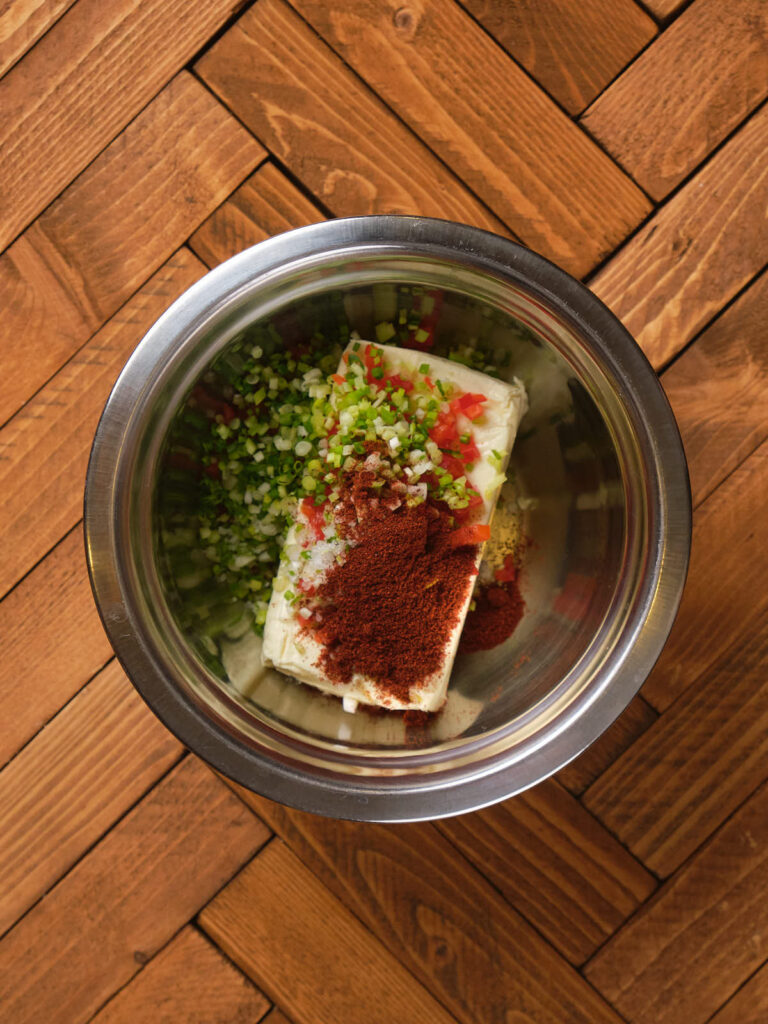 A metal bowl contains cream cheese, chopped green onions, diced tomatoes, and chili powder.