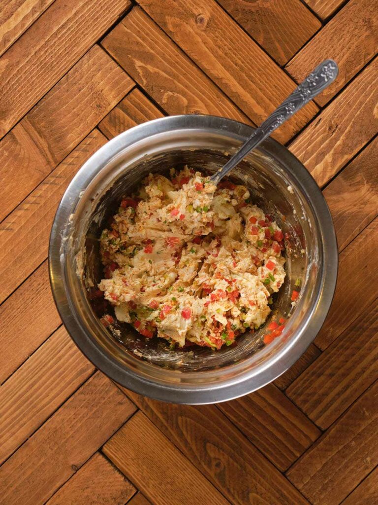 A stainless steel bowl on a wooden surface contains a mixture of chopped vegetables and mashed ingredients.